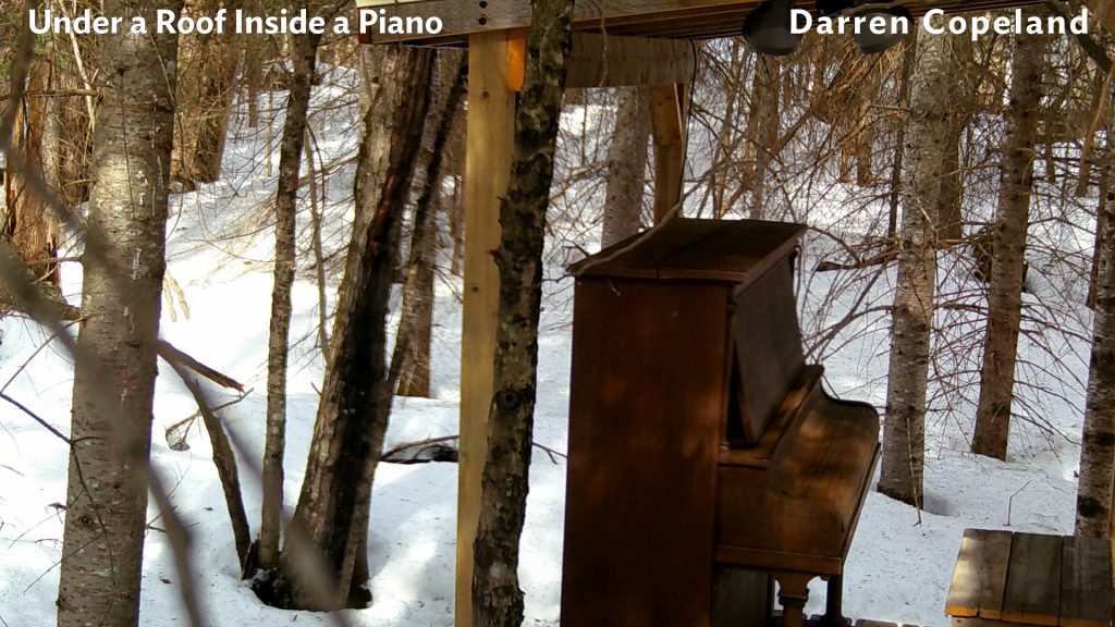 Upright piano outdoors on a deck under a shed roof. Snow covers the ground in the daytime.
